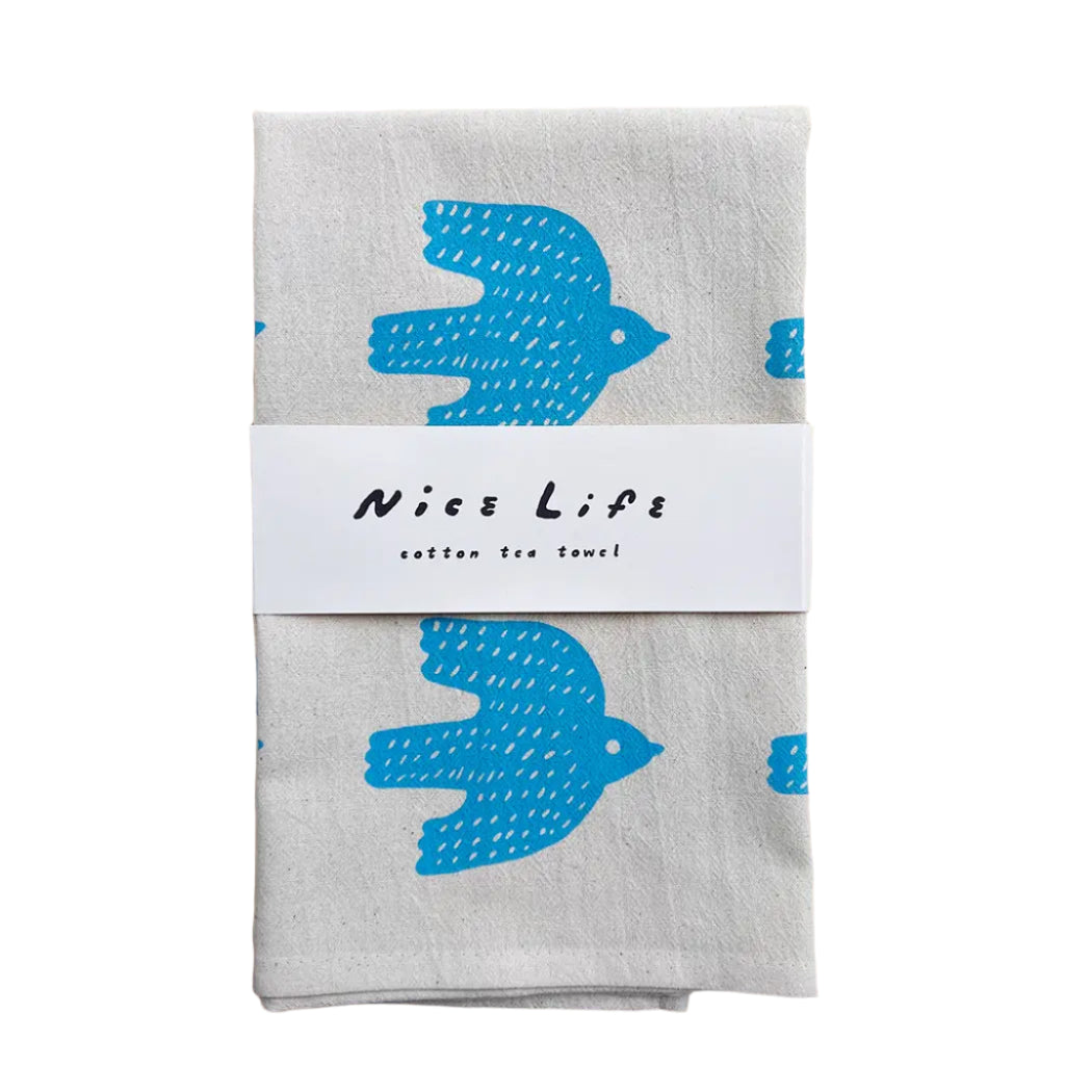 Folded tea towel with blue bird design and 'Nice Life' label on a wooden surface