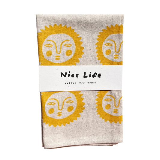 Folded tea towel with yellow face design and 'Nice Life' label on a wooden surface
